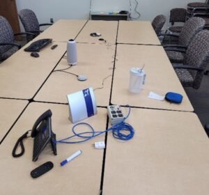 9 conference room tables moved together in a 3x3  grid.  On the top of the tables is the portable hearing loop, phone, marrkers, microphones, water bottle, etc.