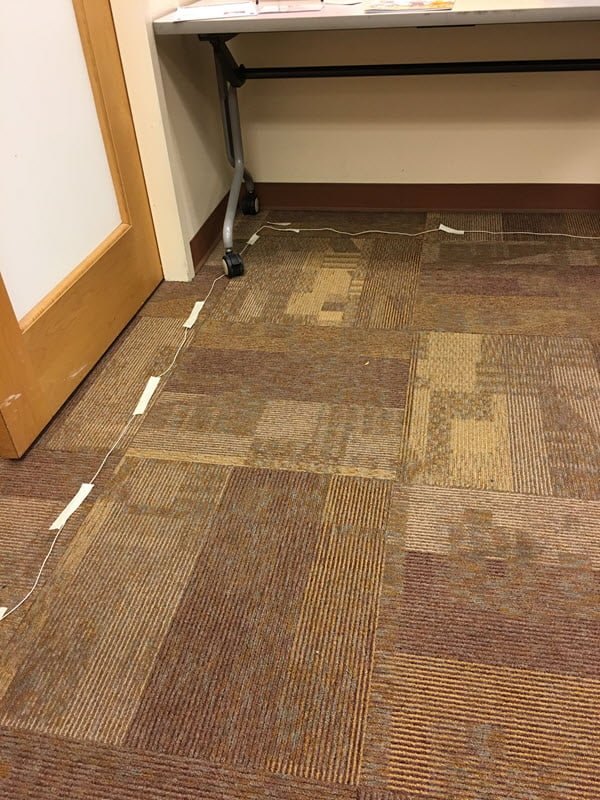 white wire on top of carpet in corner of room, with masking tape holding it down - temporary hearing loop