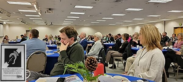 A large conference room with people sitting around round tables. An assistive listening system sign is added in the lower left corner